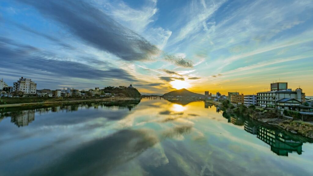 夕暮れ時の穏やかな木曽川に空の色が反射する水面と、遠方の山の上に佇む犬山城のシルエット