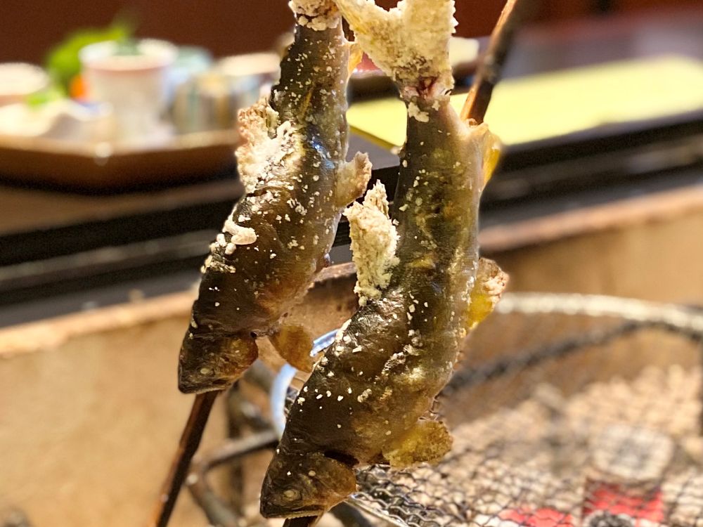 囲炉裏の炭火で串焼きにされている、塩を振った2匹の鮎。