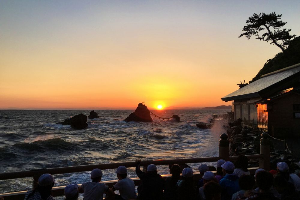 二見浦の夜明けの風景。海上の夫婦岩の間から、鮮やかなオレンジ色の太陽が昇り始めています。朝日に照らされて波しぶきが黄金色に輝き、手前の展望台では多くの参拝客や観光客がその神々しい瞬間を見守っています。