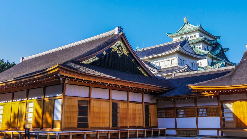 青空の下に佇む、復元された名古屋城本丸御殿の外観。背後には名古屋城の天守閣がそびえ立っている。