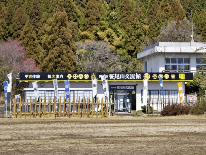 豊かな緑を背景にした笹尾山交流館の外観。白と黒のモダンな外装の平屋建ての建物で、入り口には武将の家紋が並び、前には木製の馬防柵が再現されている様子。