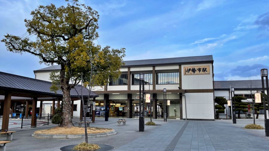 青空を背景にした伊勢市駅の駅舎外観。手前には大きな木と、外宮参道へ続く広々とした歩行者用スペースが広がっている。