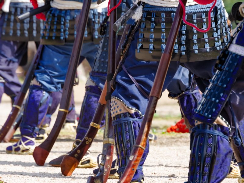 関ケ原合戦祭りに参加する甲冑武者たちの足元のカット。青い着物に脛当てを着け、草鞋（わらじ）を履いた複数の武者たちが火縄銃を持って並んでいる様子。