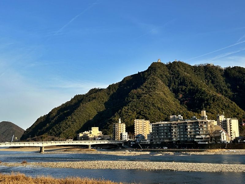 青空の下、穏やかに流れる長良川と、その背後にそびえる緑豊かな金華山。山頂には岐阜城が見え、川沿いには長良川温泉の旅館やホテルが立ち並ぶ風景。