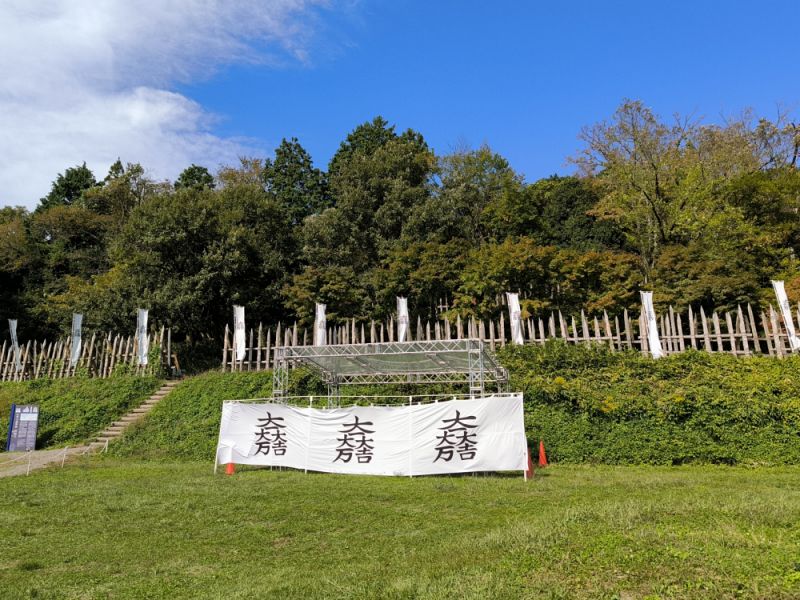 笹尾山のふもとの斜面に沿って設置された木製の馬防柵と、その手前の広場に掲げられた石田三成の紋章「大一大万大吉」が入った白い横幕。