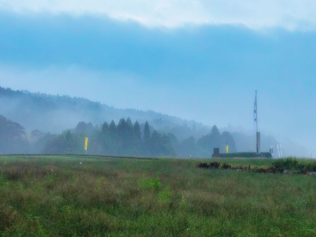 早朝の深い霧に包まれた関ケ原古戦場の草原。遠くの山々や木々が白く霞み、平地にはわずかに幟（のぼり）が見える幻想的で静かな風景。