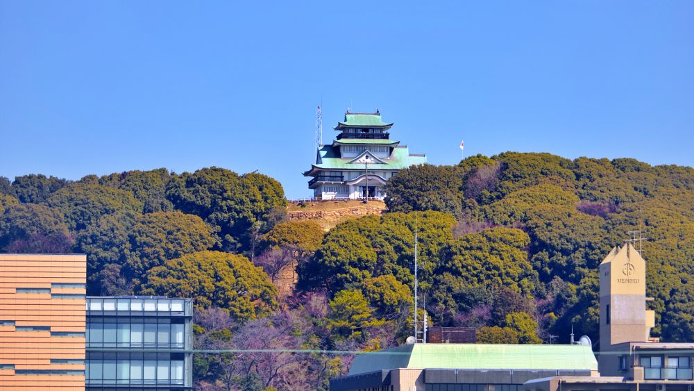 青空の下、豊かな緑に包まれた小牧山の山頂に立つ、白壁と緑の屋根が特徴的な三層四階の小牧山歴史館(城郭風建築)。