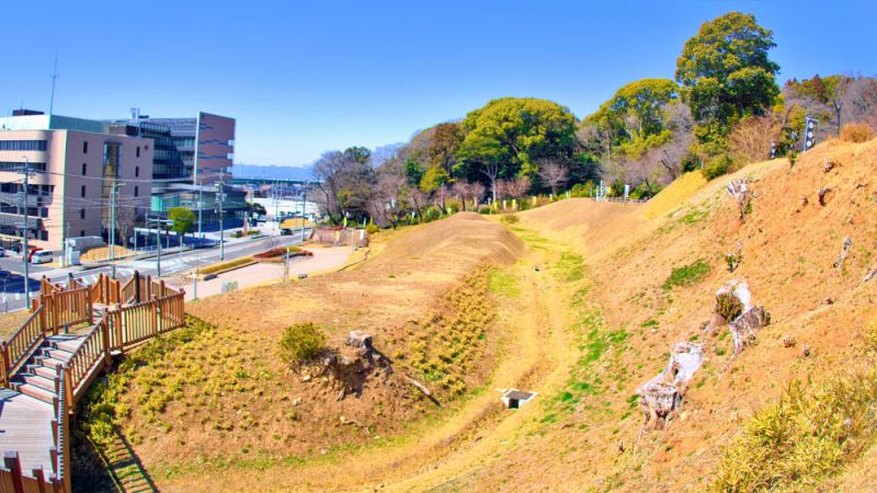 小牧山城の史跡公園内に整備された、深く大きな堀と高く盛られた土塁の遺構。手前には見学用の木製階段があり、奥には現代の街並みが広がっています。