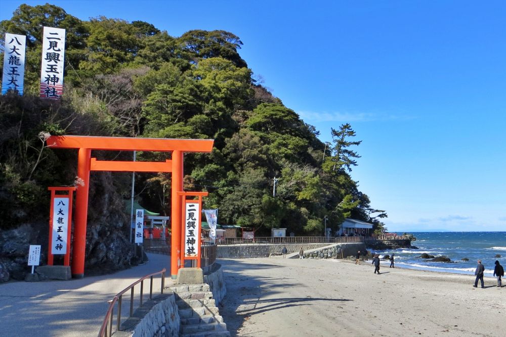 快晴の二見興玉神社。砂浜の入り口に鮮やかな朱色の鳥居が立っており、傍らには「二見興玉神社」と書かれた石柱があります。背景には緑豊かな木々と青い空が広がり、海岸沿いを数人の観光客が歩いている穏やかな風景です。