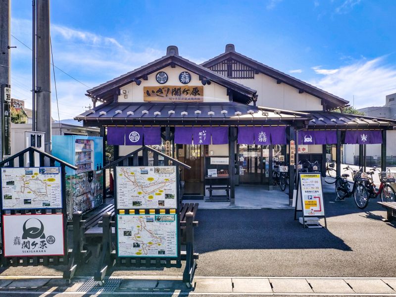 JR関ヶ原駅前にある、紫色の暖簾（のれん）が掛かった和風な外観の関ヶ原駅前観光交流館。入り口前には観光マップの看板が設置されている様子。