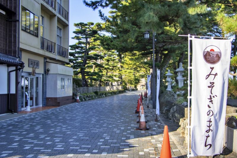 レトロな建物と松の木が並ぶ石畳の参道。手前には「みそぎのまち」と書かれた白い幟が立っている。