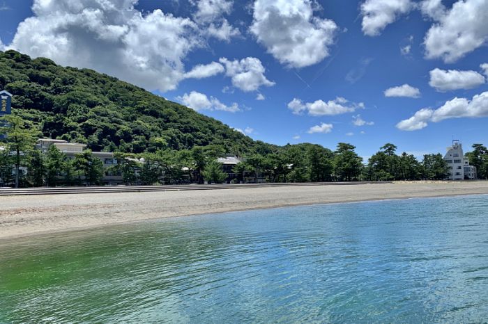 青い空と白い雲の下に広がる、穏やかで透明なエメラルドグリーンの二見浦の海と砂浜。