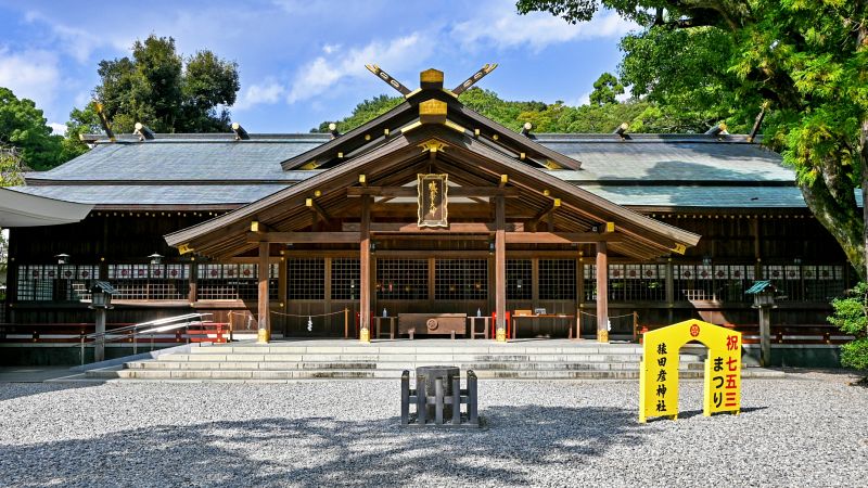 玉砂利が敷き詰められた境内に建つ、重厚な木造の猿田彦神社社殿。屋根には黄金の装飾が輝いています。