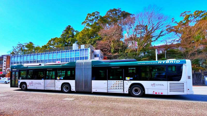青い空の下、停留所に停まっている三重交通の白と紺色の大きな連節バス。