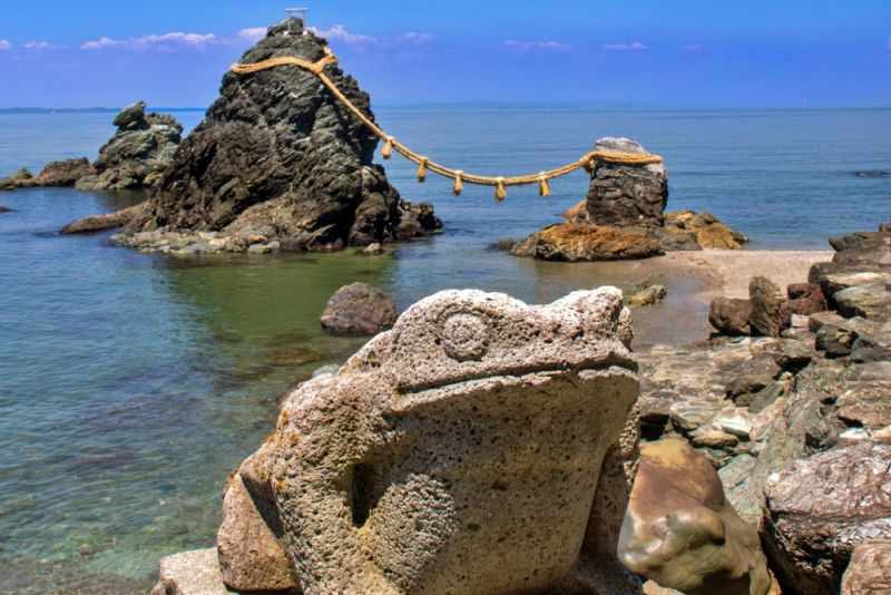 手前に鎮座するカエルの石像越しに、青い海に浮かぶ夫婦岩を望む風景。