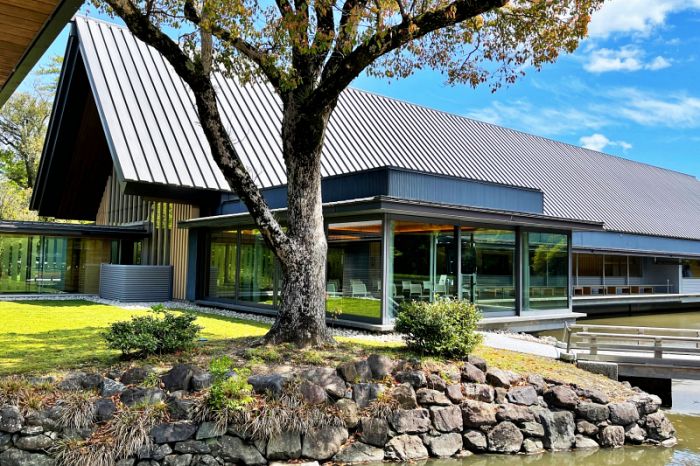 池のほとりに建つ、大きな三角屋根が特徴的なモダンな外観の博物館。手前には丁寧に手入れされた石組みと樹木があります。